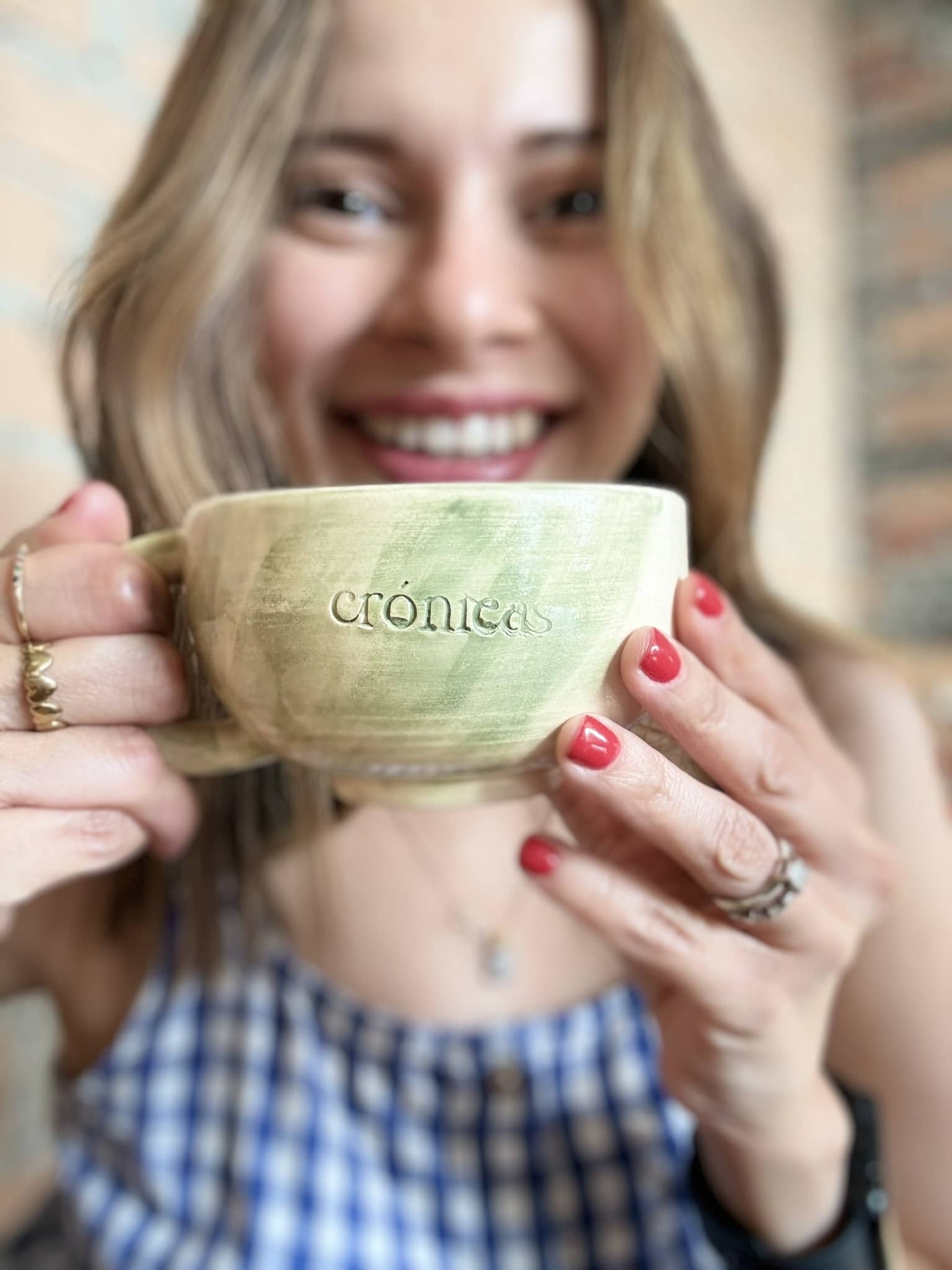 Gina holding a green cup with the word 'cronicas' engraved, wearing a blue checkered dress and red nail polish.